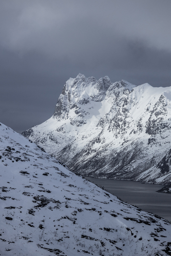 View of steep snow covered mountain peaks towering over the dark waters of a fjord under a heavy overcast winter sky Ersfjord Troms Norway. View of steep snow covered mountain peaks towering over the dark waters of a fjord under a heavy overcast winter sky Ersfjord Troms Norway.