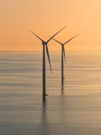 Aerial view of two offshore wind turbines standing in the calm sea under a golden sunset sky off the coast of the Netherlands.