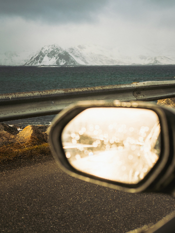 View of snow capped mountains reflecting in a car s side mirror juxtaposed against the dark ocean water under a cloudy sky Henningsv&aelig;r Norway.