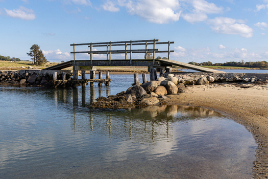 View of a wooden footbridge crossing a narrow inlet with rocky banks and sandy shores on the island of Hjarno in Hedensted Municipality Denmark. View of a wooden footbridge crossing a narrow inlet with rocky banks and sandy shores on the island of Hjarno in Hedensted Municipality Denmark.