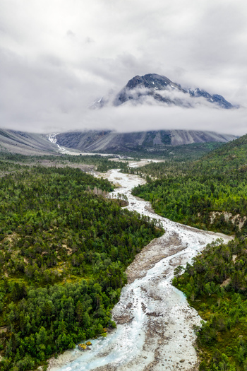 Aerial view of Faueldalen valley featuring a glacial river flowing through dense pine forests towards cloud covered mountains in Jovik Troms Norway.