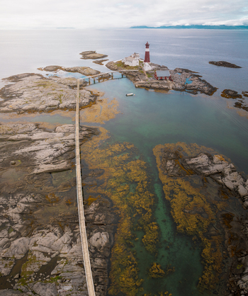 Aerial view of Tranoy Lighthouse situated on a rocky island connected by a long wooden footbridge to the shore surrounded by clear turquoise waters and seaweed in Tranoy Nordland Norway.