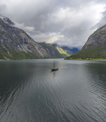 Aerial view of a traditional wooden sailing boat on the calm waters of Hardangerfjord surrounded by steep rocky mountains and green slopes in Blurnes Vestland Norway. Aerial view of a traditional wooden sailing boat on the calm waters of Hardangerfjord surrounded by steep rocky mountains and green slopes in Blurnes Vestland Norway.