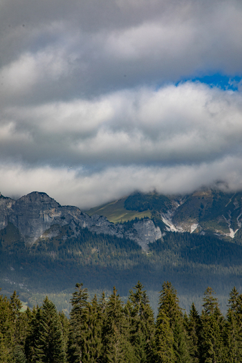View of the Swiss Alps with dramatic clouds hanging over the rugged mountain peaks and a dense pine forest in the foreground in Airolo Ticino Switzerland. View of the Swiss Alps with dramatic clouds hanging over the rugged mountain peaks and a dense pine forest in the foreground in Airolo Ticino Switzerland.