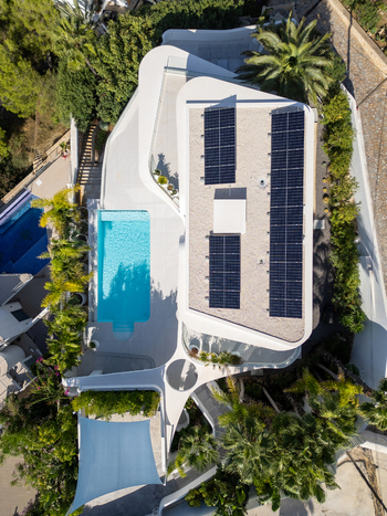 Aerial view of a modern white villa with a swimming pool and solar panels on the roof surrounded by palm trees and lush greenery in Moraira Valencian Community Spain.