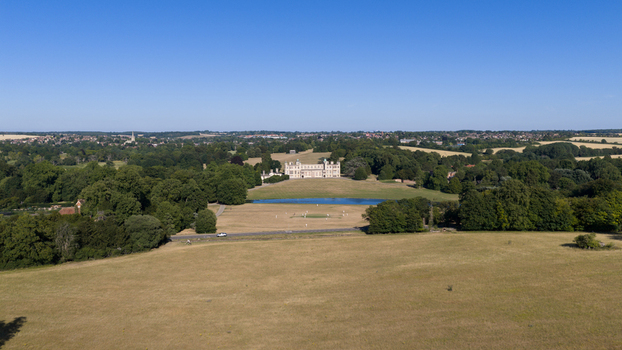 Aerial view of Audley End House surrounded by expansive green parkland a small lake and mature trees under a clear blue sky in Audley End England United Kingdom. Aerial view of Audley End House surrounded by expansive green parkland a small lake and mature trees under a clear blue sky in Audley End England United Kingdom.