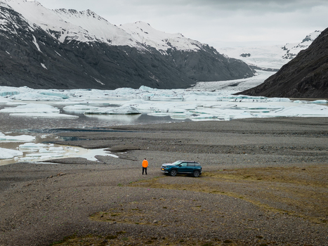 Aerial view of Vatnajokull glacier with a person in an orange jacket standing near a car by the glacial lake filled with icebergs under a cloudy sky in Iceland.