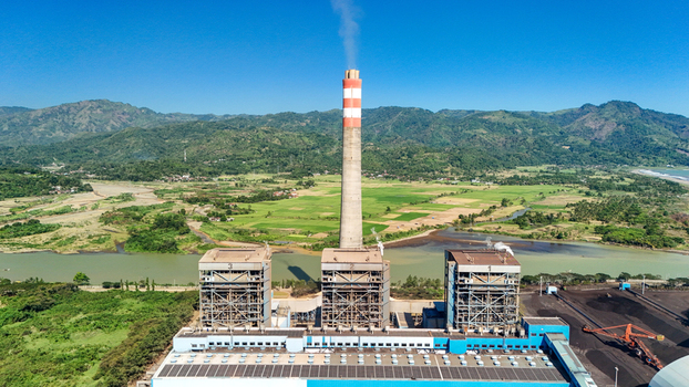 Aerial view of a power plant with a tall chimney and industrial buildings situated near a river and lush green mountains in Indonesia.