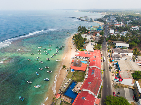 Aerial view of the turquoise coastline with anchored boats sandy beaches and urban buildings along the shore in Hikkaduwa Southern Province Sri Lanka. Aerial view of the turquoise coastline with anchored boats sandy beaches and urban buildings along the shore in Hikkaduwa Southern Province Sri Lanka.