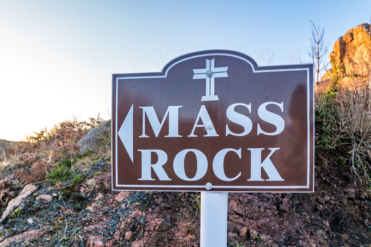 View of a brown directional sign for the historic Kincasslagh Mass Rock set against a rocky landscape during golden hour in Kincasslagh County Donegal Ireland. View of a brown directional sign for the historic Kincasslagh Mass Rock set against a rocky landscape during golden hour in Kincasslagh County Donegal Ireland.