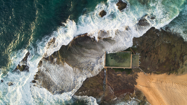 Aerial view of Newport Rock Pool with waves crashing against rugged rocks and people swimming during golden hour in Sydney New South Wales Australia.