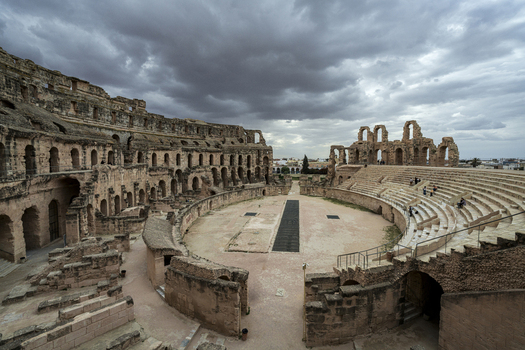 View of the ancient Roman Amphitheatre of El Jem with its weathered stone arches tiered seating and dramatic cloudy sky in El Jem Gouvernorat de Mahdia Tunisia.