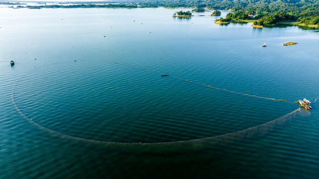 Aerial view of a large fishing net being pulled by a boat in the calm waters of Kaptai Lake surrounded by lush green hills and small islands Rangamati Chittagong Division Bangladesh.