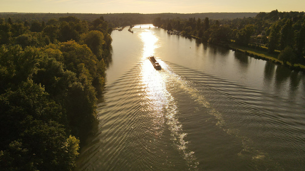 Aerial view of a barge cruising along the Seine river at sunset creating a wake in the golden light surrounded by lush green trees in Chartrettes Ile de France France. Aerial view of a barge cruising along the Seine river at sunset creating a wake in the golden light surrounded by lush green trees in Chartrettes Ile de France France.