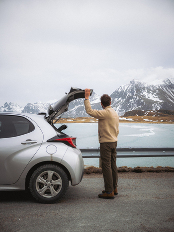 View of a man at the back of his silver car gazing toward the serene lake and the majestic snow dusted mountains Fredvang Nordland Norway. View of a man at the back of his silver car gazing toward the serene lake and the majestic snow dusted mountains Fredvang Nordland Norway.
