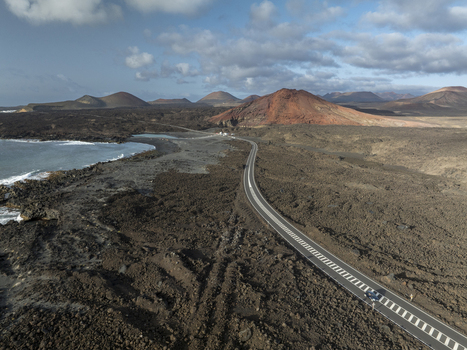 Aerial view of a winding road cutting through rugged volcanic lava fields with distant craters and the Atlantic Ocean coastline in Lanzarote Canarias Spain. Aerial view of a winding road cutting through rugged volcanic lava fields with distant craters and the Atlantic Ocean coastline in Lanzarote Canarias Spain.