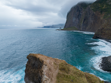 Aerial view of a rugged coastal cliff overlooking the deep blue Atlantic Ocean with crashing waves and a dramatic cloudy sky in Madeira. Aerial view of a rugged coastal cliff overlooking the deep blue Atlantic Ocean with crashing waves and a dramatic cloudy sky in Madeira.