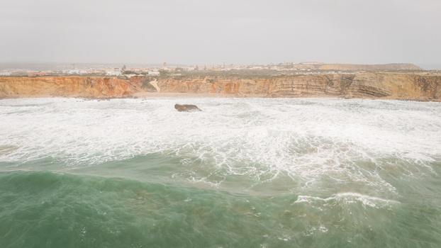 Aerial view of rough turquoise waves crashing against the rugged orange cliffs of the coastline near the town under an overcast sky Sagres Faro Portugal. Aerial view of rough turquoise waves crashing against the rugged orange cliffs of the coastline near the town under an overcast sky Sagres Faro Portugal.