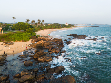 Aerial view of the rocky coastline and sandy beach with crashing waves palm trees and people walking along the shore in Galle Southern Province Sri Lanka. Aerial view of the rocky coastline and sandy beach with crashing waves palm trees and people walking along the shore in Galle Southern Province Sri Lanka.