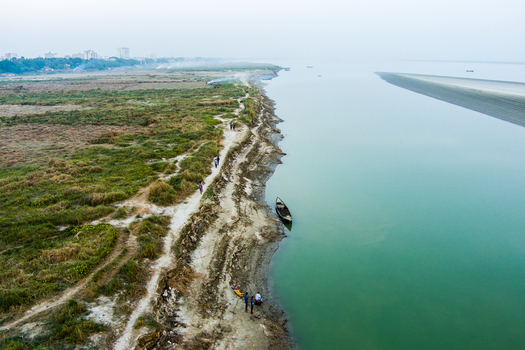 Aerial view of the Padma River bank with people walking along a dirt path a small wooden boat moored at the edge and the distant city skyline in Rajshahi Rajshahi Division Bangladesh.