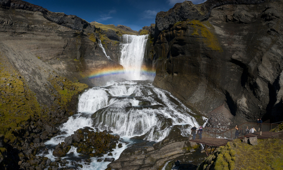 Aerial view of Dynjandi waterfall cascading down dark volcanic cliffs with a vibrant rainbow and tourists on a viewing platform in Iceland.