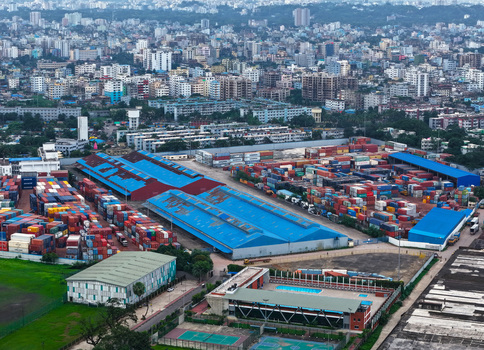 Aerial view of Chattogram Port featuring colorful shipping containers and industrial warehouses with blue roofs set against the urban skyline of Chittagong Chittagong Division Bangladesh.