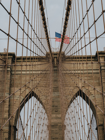 View of the Brooklyn Bridge with its iconic stone arches steel suspension cables and an American flag waving against a cloudy sky in New York New York United States.