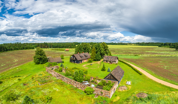 Aerial view of a rural farmstead with historic stone ruins traditional wooden houses and lush green fields under a dramatic cloudy sky in Latvia. Aerial view of a rural farmstead with historic stone ruins traditional wooden houses and lush green fields under a dramatic cloudy sky in Latvia.