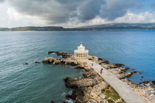 Aerial view of the Saint Theodore Lighthouse situated on a rocky peninsula surrounded by the Ionian Sea with people walking along the concrete pier under a cloudy sky in Argostoli Greece.