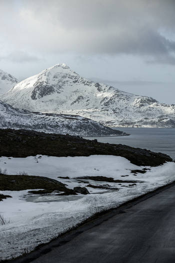 View of snow capped mountains overlooking a fjord under a cloudy sky from a roadside Kvaloya Troms Norway. View of snow capped mountains overlooking a fjord under a cloudy sky from a roadside Kvaloya Troms Norway.