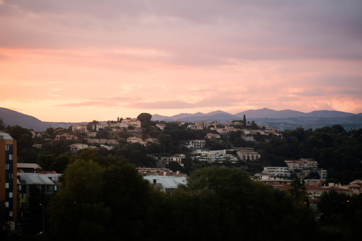 View of the hillside residential area at sunset featuring houses nestled among trees with distant mountains under a soft pink sky in Nice Provence Alpes Cote d Azur France. View of the hillside residential area at sunset featuring houses nestled among trees with distant mountains under a soft pink sky in Nice Provence Alpes Cote d Azur France.