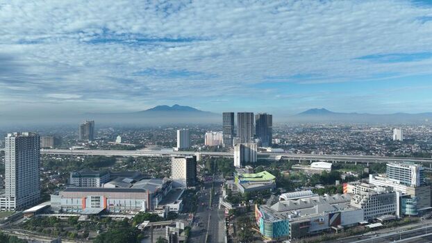 Bekasi Barat Indonesia 12 April 2026 Aerial view of the cityscape featuring modern high rise buildings a highway overpass and distant mountains under a blue sky with clouds. Bekasi Barat Indonesia 12 April 2026 Aerial view of the cityscape featuring modern high rise buildings a highway overpass and distant mountains under a blue sky with clouds.