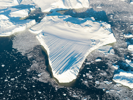 Aerial view of the Ilulissat glacier iceberg floating in the deep blue arctic waters with scattered ice fragments and bright sunlight casting shadows on the ice surface in Greenland. Aerial view of the Ilulissat glacier iceberg floating in the deep blue arctic waters with scattered ice fragments and bright sunlight casting shadows on the ice surface in Greenland.