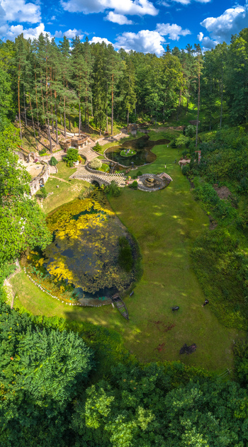 Aerial view of a lush park featuring ponds stone bridges walking paths and dense forest surroundings under a blue sky in Latvia. Aerial view of a lush park featuring ponds stone bridges walking paths and dense forest surroundings under a blue sky in Latvia.