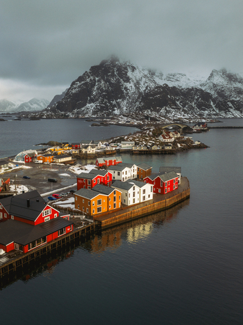 Aerial view of colorful wooden buildings nestle along the water s edge a vibrant contrast against the snow dusted mountains and the serene reflective sea Henningsv&aelig;r Nordland Norway.