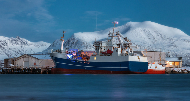 View of a large blue and white fishing vessel docked at a harbor with snow covered mountains in the background at twilight in Sommaroy Troms Norway. View of a large blue and white fishing vessel docked at a harbor with snow covered mountains in the background at twilight in Sommaroy Troms Norway.