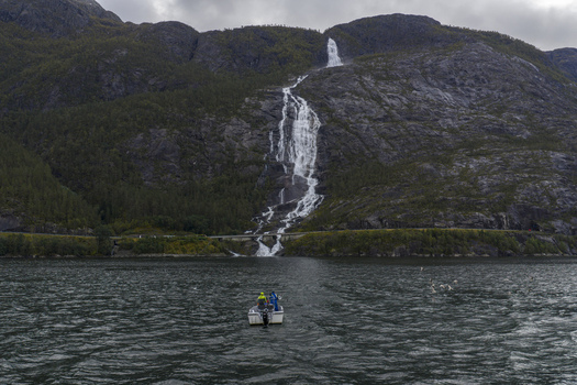 Aerial view of Langfossen waterfall cascading down steep rocky cliffs into the Akrafjorden with a small boat in the foreground in Fjaera Vestland Norway. Aerial view of Langfossen waterfall cascading down steep rocky cliffs into the Akrafjorden with a small boat in the foreground in Fjaera Vestland Norway.