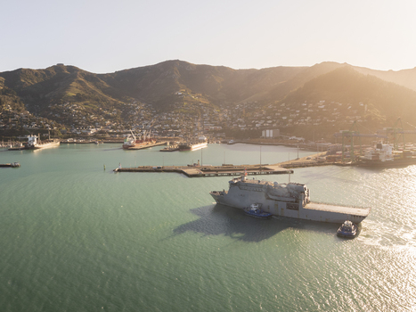 Aerial view of Lyttelton Harbour with a naval vessel being assisted by tugboats surrounded by coastal hills and port infrastructure in Christchurch Canterbury Region New Zealand.