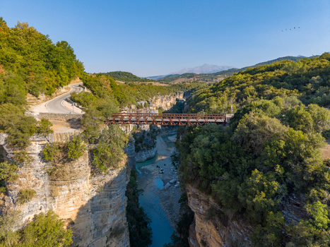 Aerial view of Osumi Canyon featuring a metal bridge spanning the deep gorge with winding roads and lush green vegetation under a clear blue sky in Dhores Berat County Albania.