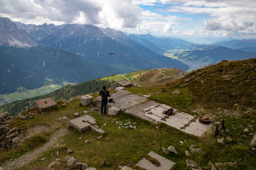 View of a person standing near historic concrete ruins on a mountain ridge overlooking the Dolomites valley with a drone in the sky Rocca Pietore Veneto Italy. View of a person standing near historic concrete ruins on a mountain ridge overlooking the Dolomites valley with a drone in the sky Rocca Pietore Veneto Italy.