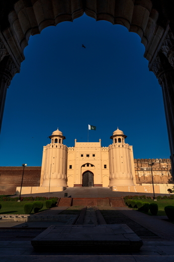 View of the historic Lahore Fort with its iconic Alamgiri Gate stone walls and a flag flying under a clear blue sky framed by a large archway in Lahore Punjab Pakistan.