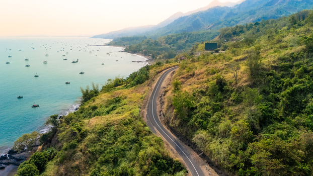Aerial view of a winding coastal road along a lush green hillside overlooking the ocean filled with small fishing boats in Sukabumi Indonesia. Aerial view of a winding coastal road along a lush green hillside overlooking the ocean filled with small fishing boats in Sukabumi Indonesia.