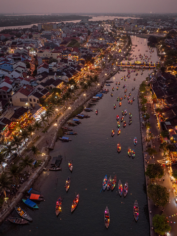 Aerial view of the Thu Bon River in Hoi An at dusk featuring numerous traditional wooden boats with glowing lanterns historic yellow buildings and palm trees lining the riverbanks in Vietnam. Aerial view of the Thu Bon River in Hoi An at dusk featuring numerous traditional wooden boats with glowing lanterns historic yellow buildings and palm trees lining the riverbanks in Vietnam.