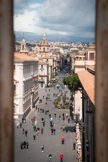 View of crowds strolling along sun drenched streets framed by weathered stone reaching toward ancient domes under a sky of swirling clouds Taormina Sicily Italy. View of crowds strolling along sun drenched streets framed by weathered stone reaching toward ancient domes under a sky of swirling clouds Taormina Sicily Italy.