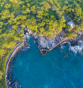 Aerial view of Mahukona Beach Park featuring a small harbor with turquoise water rocky shores and lush green trees under bright sunlight Waimea Hawaii United States.