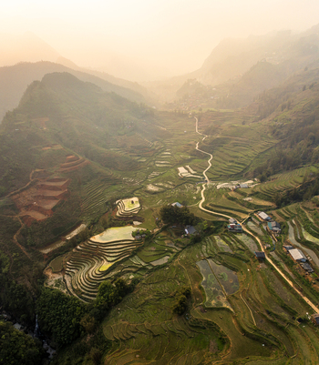Aerial view of lush green terraced rice fields carved into the mountainside with a winding path and small rural houses under a soft golden sunset light in Vietnam. Aerial view of lush green terraced rice fields carved into the mountainside with a winding path and small rural houses under a soft golden sunset light in Vietnam.