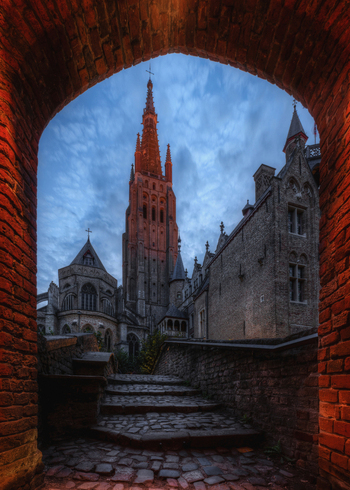 View of the Church of Our Lady framed by a historic brick archway with cobblestone steps leading towards the gothic tower in Bruges Flanders Belgium.