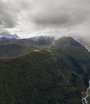 Aerial view of Buarbreen glacier and lush green mountain slopes with a small turquoise lake and a waterfall under a cloudy sky Ullensvang Vestland Norway. Aerial view of Buarbreen glacier and lush green mountain slopes with a small turquoise lake and a waterfall under a cloudy sky Ullensvang Vestland Norway.
