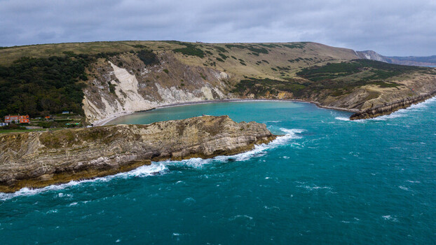 Aerial view of Lulworth Cove with turquoise water dramatic limestone cliffs and rolling green hills along the coast of the United Kingdom.