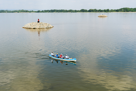View of people paddling a blue canoe on calm water past small stone islands and a distant green shoreline in Bratislava Bratislava Region Slovakia. View of people paddling a blue canoe on calm water past small stone islands and a distant green shoreline in Bratislava Bratislava Region Slovakia.